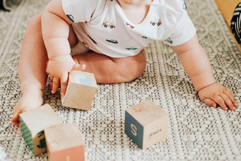 A baby sitting on the floor in front of some wooden blocks. It head is out of frame but its posture suggests movement, as if about to  begin to reach or crawl to someone also out of frame.