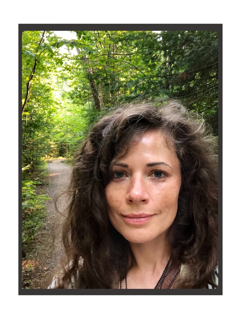 A woman with long hair smiles contentedly into the camera. Behind her a trail leads into a lush forest of broadleaves and evergreens. 