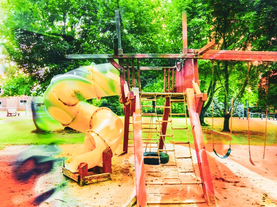 A colorful image of an outdoor play set with a slide, on a sunny day. Trees are in the background, this looks like the playground of a small park.