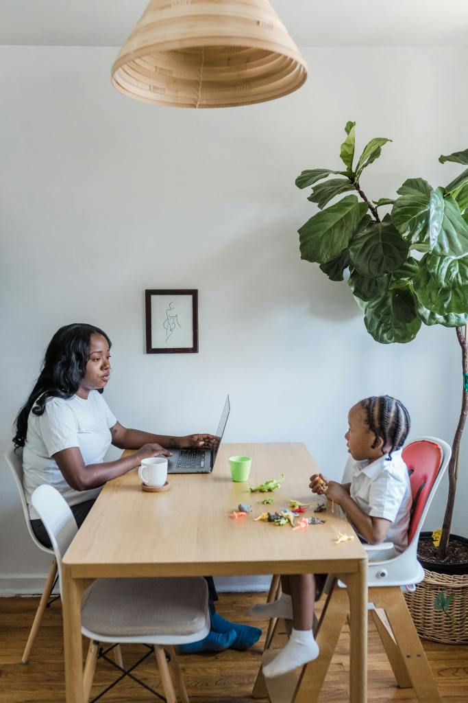 A woman and small child sit at a kitchen table looking at one another. The woman is working on a laptop and the child is in a high chair with a collection of toy dinosaurs in front of her.