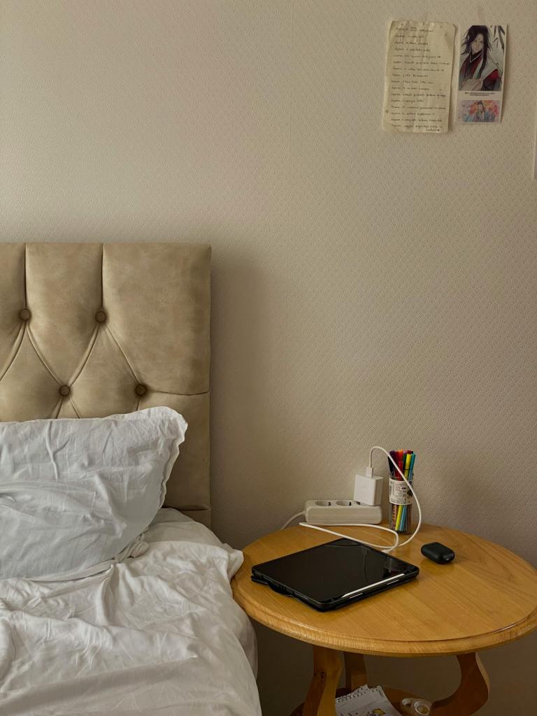 a bed in a simply decorated room. There is a laptop on the nightstand, some pens, and a case for ear buds. A note and an asian illustration are hung on the wall.